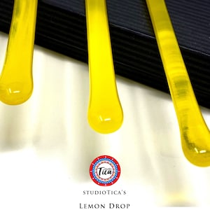 May include: Three yellow glass rods with rounded ends, dripping down from a black surface. The rods are part of a set of glass art pieces called "Lemon Drop Family" by Studio Tica.
