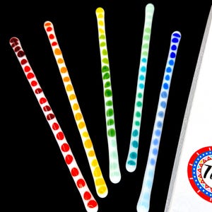 May include: Five glass stirring sticks with colourful polka dots. The sticks are arranged in a rainbow pattern, with red, orange, yellow, green, and blue dots. The sticks are on a black background.