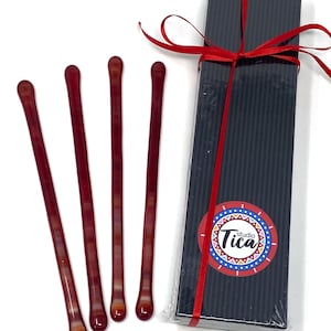 Pode incluir: Conjunto de quatro palitos de agitação de vidro vermelho em uma caixa preta com uma fita vermelha e um logotipo colorido que diz "studio Tica".