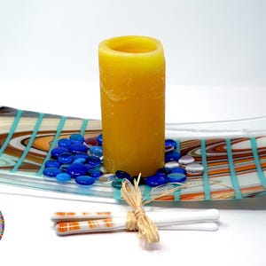 A glass tray with a Southwest pattern holding a yellow pillar candle, blue glass gems, and a pair of white decorative chopsticks.