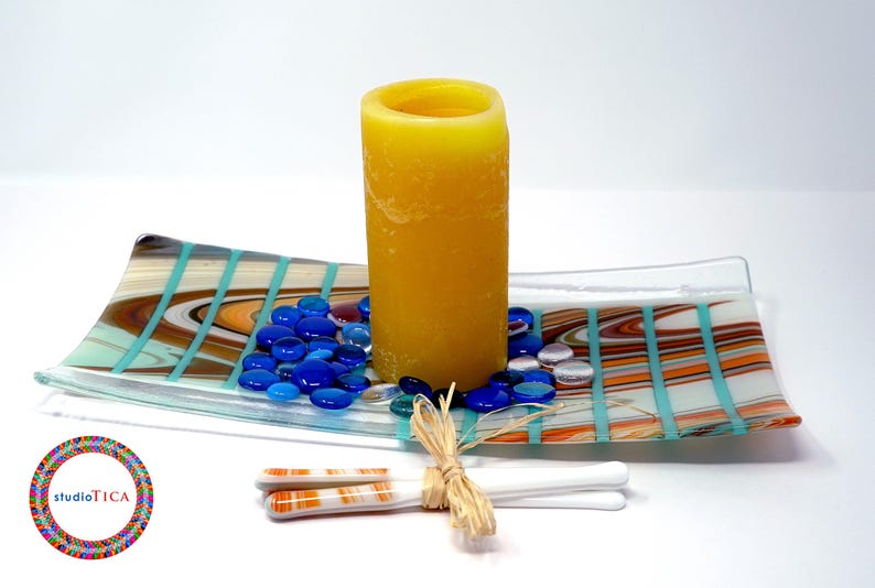 A glass tray with a Southwest pattern holding a yellow pillar candle, blue glass gems, and a pair of white decorative chopsticks.