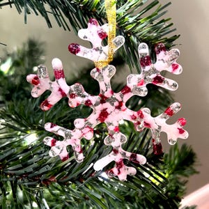 May include: A white glass snowflake ornament with red and silver glitter accents. The snowflake is hanging on a Christmas tree.