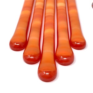 May include: Five orange glass sticks with a gradient of color from light orange to dark orange. The sticks are arranged in a fan shape.