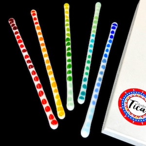 Könnte beinhalten: Sechs Glasrührstäbchen mit weißem Hintergrund und bunten Punkten. Die Stäbchen sind in einem Regenbogenmuster angeordnet, mit roten, orangefarbenen, gelben, grünen, blauen und violetten Punkten.