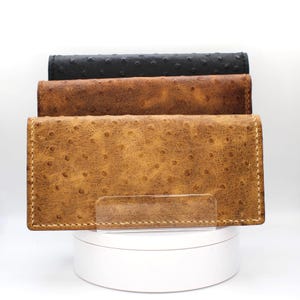 May include: Three rectangular wallets stacked vertically, featuring a textured ostrich-style pattern. The top wallet is black, the middle is a medium brown, and the front wallet is a lighter tan, all finished with visible light-coloured stitching.