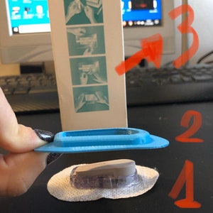 Op de afbeelding: Een close-up van een transparant medisch apparaat met een grijze bovenkant, dat op een wit gaasje ligt. Het apparaat is omgeven door een lichtblauwe plastic ring. De afbeelding toont ook een doos 3M Tegaderm Roll 16004S.