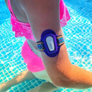 Op de afbeelding: Een waterdichte polsband in paars en wit met een kleurrijk kralenband. De polsband wordt gedragen op de arm van een persoon in een zwembad.