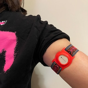 May include: A red and black patterned arm band with a small, round, red device attached. The device has the text "nock.com" printed on it.