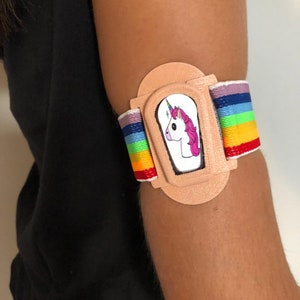 Puede incluir: Un brazalete de tela beige con un diseño de unicornio rosa y blanco y una banda elástica arcoíris.