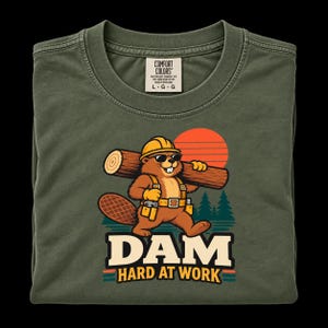 May include: An olive green t-shirt featuring a graphic of a brown beaver wearing a yellow hard hat and sunglasses, carrying a log. The beaver wears a tool belt. Below the graphic, the text reads DAM HARD AT WORK in bold, cream-coloured letters.