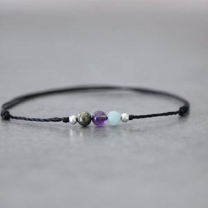 Peut inclure: Un bracelet en cordon noir avec trois perles : une perle dorée, une perle d'améthyste violette et une perle d'aigue-marine bleu clair.