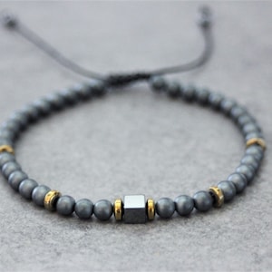 Peut inclure: Un bracelet gris perlé avec des accents dorés et une perle carrée grise au centre. Le bracelet a un cordon noir pour ajuster la taille.