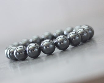 Puede incluir: Una pulsera de cuentas de color gris oscuro. Las cuentas esféricas son lisas y muy reflectantes, creando una apariencia brillante. La pulsera está ensartada, formando una forma circular. Las cuentas parecen estar hechas de un material denso.