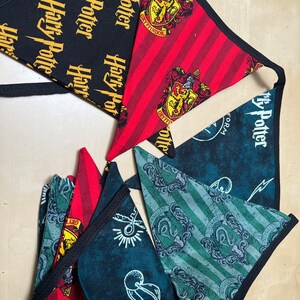 Op de afbeelding: Een set van vijf Harry Potter-thema vlaggetjes van stof. De vlaggetjes zijn driehoekig en hebben verschillende ontwerpen, waaronder de wapenschilden van de Hogwarts-huizen, het Harry Potter-logo en andere Harry Potter-beelden. De vlaggetjes zijn zwart, rood en groen.