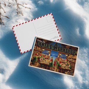 May include: Two postcards on a snowy surface. One postcard features a festive illustration of Santa's Workshop with elves. The other postcard has a blank white surface with red and white trim. The text "Greetings from Santa's Workshop" is visible.