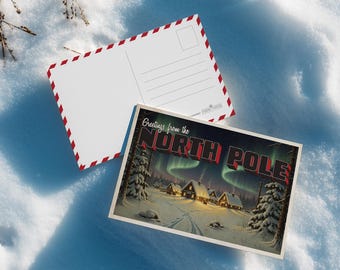 May include: Two postcards on a snowy surface. One postcard features a vintage illustration of a snowy village under the Northern Lights with the text "Greetings from the NORTH POLE". The other postcard is blank with a red and white border.