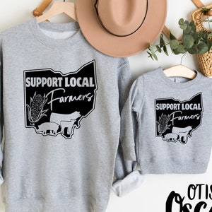 May include: Two gray sweatshirts with the text "Support Local Farmers" in black lettering inside a black outline of the state of Ohio. The design also includes a corn stalk, pig, and cow. The smaller sweatshirt has the names "Otis & Oscar" in black cursive lettering below it.