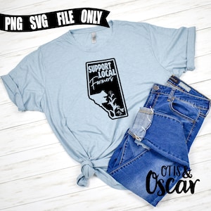 Op de afbeelding: Een lichtblauw T-shirt met een zwarte afbeelding waarop in een gestileerd lettertype "Support Local Farmers" staat. De afbeelding bevat ook een silhouet van een koe en een ma&iuml;skolf. Het T-shirt is in een knoop geknoopt en ligt op een houten oppervlak met een blauwe spijkerbroek.