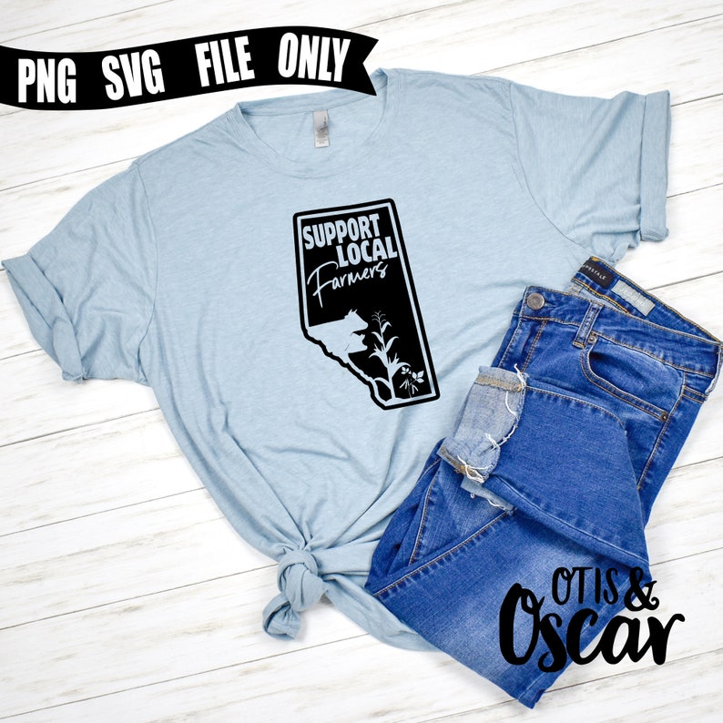Op de afbeelding: Een lichtblauw T-shirt met een zwarte afbeelding waarop in een gestileerd lettertype "Support Local Farmers" staat. De afbeelding bevat ook een silhouet van een koe en een ma&iuml;skolf. Het T-shirt is in een knoop geknoopt en ligt op een houten oppervlak met een blauwe spijkerbroek.