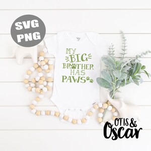 Op de afbeelding: Witte baby romper met groene tekst die "Mijn grote broer heeft pootjes" zegt met een poot afdruk grafiek. De romper ligt op een wit houten oppervlak met een witte houten kralen ketting en een groene plant.