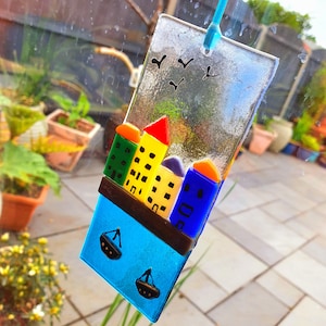May include: A colourful glass suncatcher with a cityscape design. The cityscape features buildings in shades of yellow, green, blue, and red. The suncatcher also features two small boats in the water below the cityscape. The suncatcher is hanging from a window.