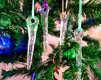 May include: Five clear glass icicle ornaments with glitter hanging on a green artificial Christmas tree. The icicles are decorated with a silver glitter finish.