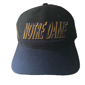 Puede incluir: Una gorra de béisbol negra y azul marino con el texto "NOTRE DAME" en letras amarillas y azules.