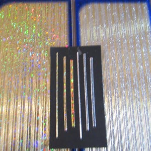 May include: Close-up of two rectangular trays with a blue border, filled with gold and silver glitter strands. A black card displays several glittery, vertical strips in various colors and widths, creating a shimmering effect.
