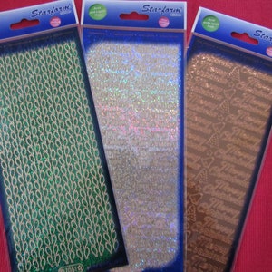 Peut inclure: Trois paquets de feuilles adhésives décoratives avec des motifs différents. La première feuille présente un motif de feuilles répétées en vert et or. La deuxième feuille présente un motif iridescent argenté. La troisième feuille présente un motif brun de sapins de Noël répétés et les mots "Joyeux Noël".