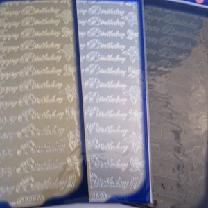 Peut inclure: Trois feuilles d'autocollants en feuille d'argent avec les mots "Joyeux anniversaire" répétés sur chaque feuille. Les feuilles sont emballées dans du plastique transparent.