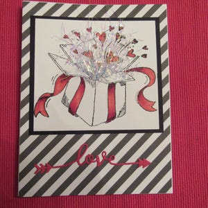 May include: A white gift box with a red ribbon and hearts bursting out of the top. The box is on a black and white striped background with the word "love" written in red.