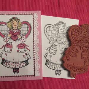 May include: A pink card with a white border and a white lace trim. The card features a black and white illustration of an angel holding a heart that says "Love". The angel is wearing a dress with a heart pattern. The card is next to a rubber stamp of the same angel.