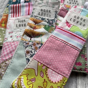 Può includere: Una collezione di segnalibri in tessuto fatti a mano con la scritta "BOOK LOVER" su ognuno. I segnalibri presentano un design patchwork con vari motivi, tra cui floreali, pois e righe. Ogni segnalibro ha un nastro bianco.