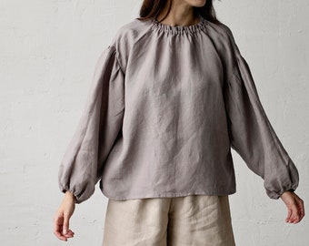 May include: A woman wearing a loose-fitting, gray linen blouse with a gathered neckline and long sleeves. She is also wearing beige linen shorts and brown leather shoes.
