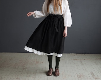 Puede incluir: Una mujer con una blusa blanca de manga larga, una falda negra y un cinturón negro. Lleva medias verdes y zapatos marrones.