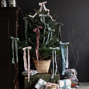 May include: A Christmas tree adorned with pastel-coloured ribbons and bows. Wrapped presents in pale blue and white paper are arranged at the base. A wicker basket supports the tree, with a vintage tin and wooden tray nearby.