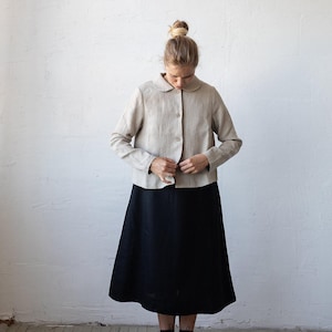 May include: A woman wearing a beige linen collared shirt with a black skirt and black lace-up boots.
