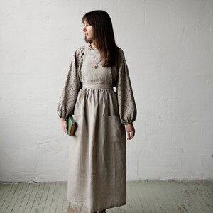 K&ouml;nnte beinhalten: Eine Frau tr&auml;gt eine lange, beige Leinen-Sch&uuml;rze mit einem floralen Stickmuster. Sie tr&auml;gt eine schwarz-wei&szlig; gepunktete Bluse und h&auml;lt ein gr&uuml;nes Buch.