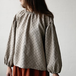 K&ouml;nnte beinhalten: Eine beige Bluse mit schwarzen Punkten und langen, gerafften &Auml;rmeln. Die Bluse wird mit einem rostfarbenen Rock kombiniert.
