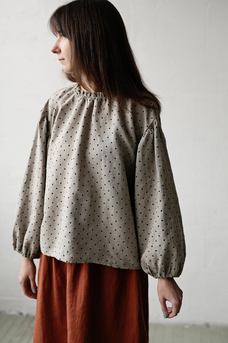 K&ouml;nnte beinhalten: Eine beige Bluse mit schwarzen Punkten und langen, gerafften &Auml;rmeln. Die Bluse wird mit einem rostfarbenen Rock kombiniert.