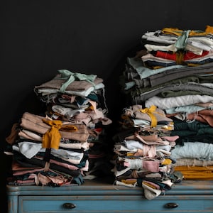 Op de afbeelding: Twee stapels stofbundels, vastgebonden met linten, in tinten beige, roze en groen, staan op een blauwe, verweerde commode. De bundels hebben verschillende maten en texturen, wat een rustieke uitstraling creëert.