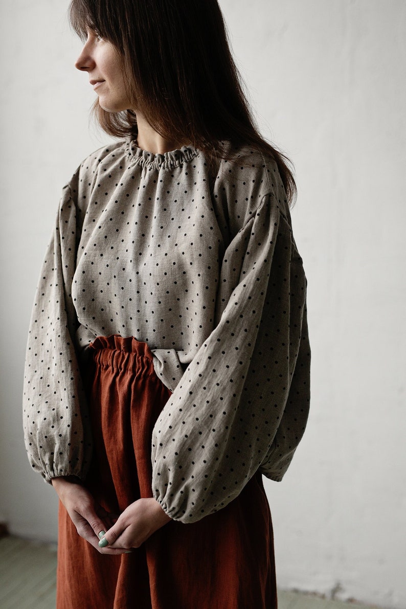 K&ouml;nnte beinhalten: Eine Frau tr&auml;gt eine beige Bluse mit schwarzen Punkten und einen rostfarbenen Rock. Die Bluse hat lange, bauschige &Auml;rmel.