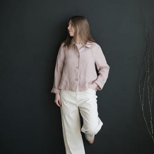 May include: A woman wearing a light pink linen shirt and white wide-leg pants. She is standing in front of a black wall.