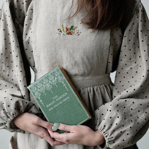 K&ouml;nnte beinhalten: Eine Frau tr&auml;gt ein hellbraunes Leinenkleid mit schwarzen Punkten und eine passende Sch&uuml;rze. Die Sch&uuml;rze ist mit einem gestickten Motiv eines Hirsches in einem Wald verziert. Sie h&auml;lt ein gr&uuml;nes Buch mit dem Titel "The Magic Garden" von Gene Stratton-Porter.
