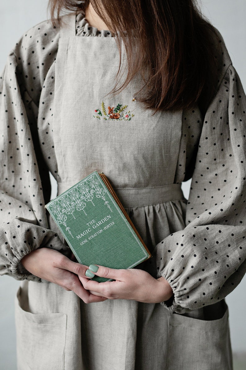 K&ouml;nnte beinhalten: Eine Frau tr&auml;gt ein hellbraunes Leinenkleid mit schwarzen Punkten und eine passende Sch&uuml;rze. Die Sch&uuml;rze ist mit einem gestickten Motiv eines Hirsches in einem Wald verziert. Sie h&auml;lt ein gr&uuml;nes Buch mit dem Titel "The Magic Garden" von Gene Stratton-Porter.