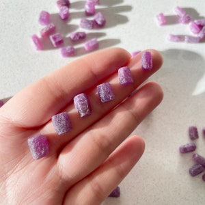 May include: A collection of small, rectangular, purple gemstones. The gemstones have a rough, textured surface and are arranged in a row on a hand.