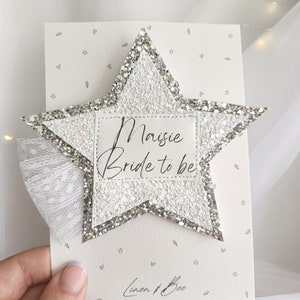 May include: A white glitter star with a silver border and a white label that says "Maisie Bride to be". The star is on a white card with a gray pattern of stars and hearts. The card also has the text "Linen & Bee" in gray.
