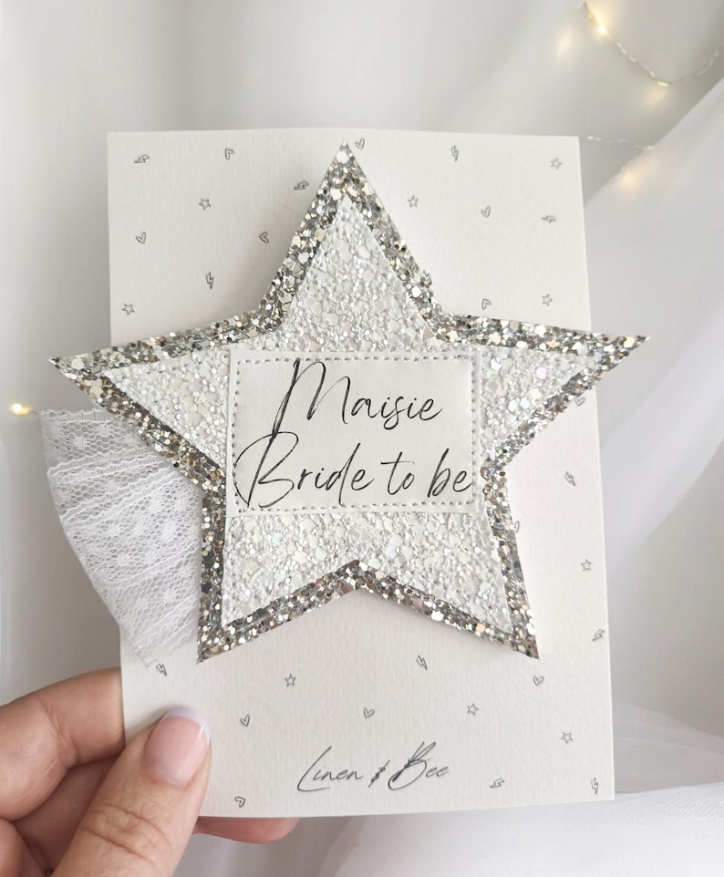 May include: A white glitter star with a silver border and a white label that says "Maisie Bride to be". The star is on a white card with a gray pattern of stars and hearts. The card also has the text "Linen & Bee" in gray.