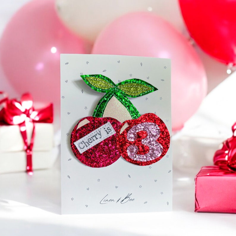 May include: A white card with a glittery red and pink cherry with a green stem and a white label that says "Cherry is 3". The card has a white background with small gray hearts and stars.