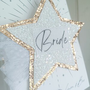 May include: A white glitter star with a gold glitter border and a white tulle bow. The star has a white label with the word "Bride" written in black cursive.